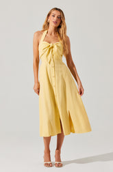 A woman is wearing a yellow sleeveless dress with a halter neckline and a large bow at the front. The dress features a button-down design running from the waist to the hem, with a slit in the front. She is also wearing open-toed beige high heel shoes.